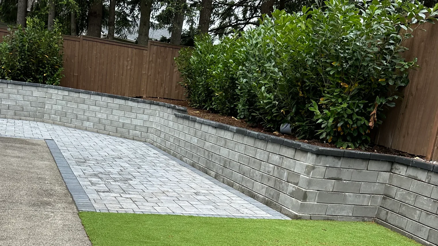 Landscaping company project: an artificial turf lawn, a paver walkway and a curved block retaining wall below a laurel hedge and cedar fence
